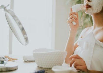 pexels-photo-8740315-8740315 A woman in white applying facial cream with a cleansing brush during her skincare routine.